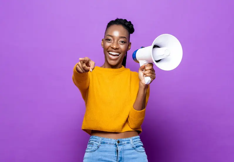 Smiling woman in a yellow sweater holding a megaphone and pointing forward, standing against a bright purple background.
