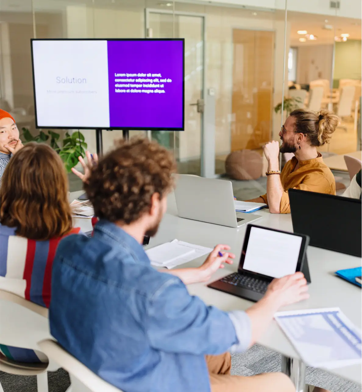 Four people sit around a conference table with laptops, discussing a presentation displayed on a screen. One person gestures toward the screen, while others listen attentively in a modern office setting.