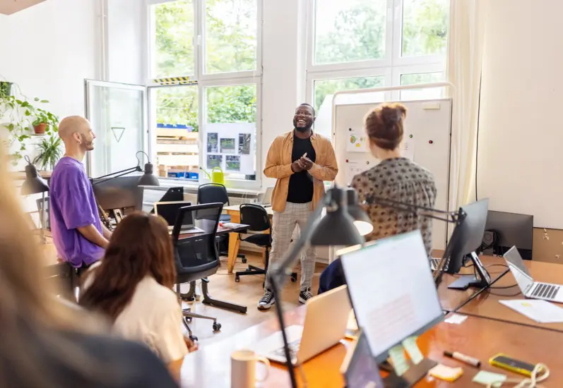 Four people in a bright, modern office are having a meeting. One person stands and speaks enthusiastically near a whiteboard, while the others sit or stand around desks with laptops and office supplies.