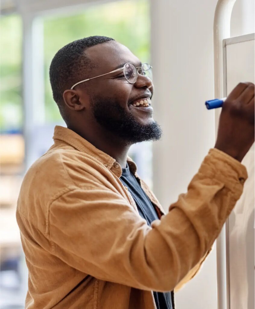 A smiling man with glasses and a beard writes on a whiteboard using a blue marker. He is wearing a tan jacket over a dark shirt and appears to be in a bright, indoor setting.