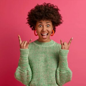 A person with curly hair, wearing a green sweater and orange hoop earrings, stands against a pink background, smiling widely and making the rock on hand gesture with both hands.