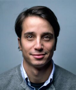 A man with short dark hair and brown eyes is smiling slightly at the camera. He is wearing a gray sweater over a white collared shirt. The background is plain and light blue.