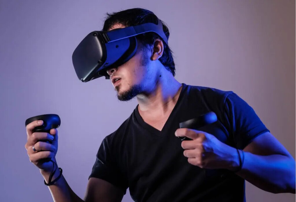 A person wearing a black virtual reality headset and holding two controllers, dressed in a dark t-shirt, appears focused while engaging with a VR experience against a plain background.