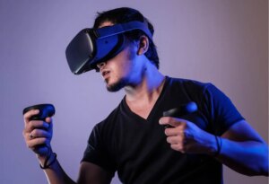 A person wearing a black virtual reality headset and holding two controllers, dressed in a dark t-shirt, appears focused while engaging with a VR experience against a plain background.