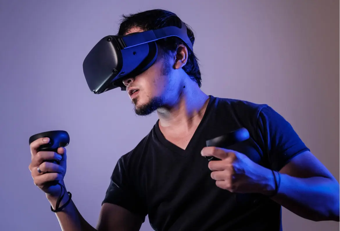 A person wearing a black virtual reality headset and holding two controllers, dressed in a dark t-shirt, appears focused while engaging with a VR experience against a plain background.