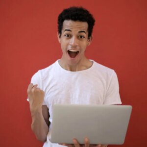 A young person in a white t-shirt stands in front of a red background, smiling excitedly with a clenched fist and holding an open laptop.