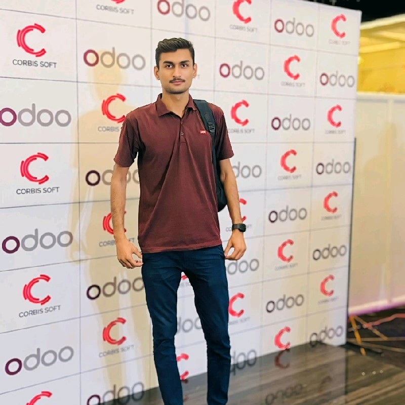 A man standing in front of a branded backdrop featuring Odoo logos at a promotional event. He is wearing a maroon polo shirt and blue jeans, with a backpack slung over one shoulder.